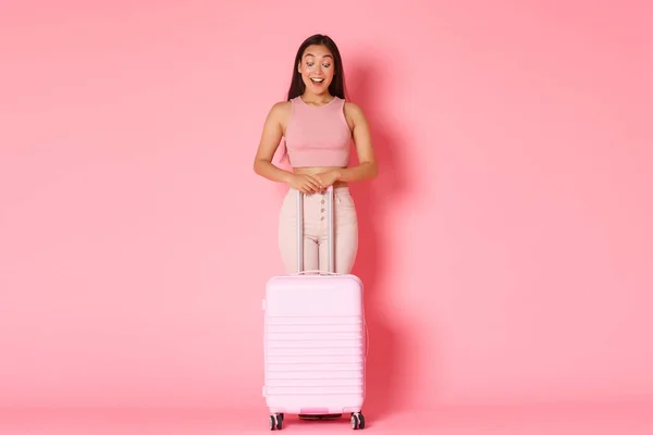 Reisen, Urlaub und Urlaubskonzept. In voller Länge aufgeregt und beeindruckt asiatische attraktive Mädchen, die mit erstaunt glückliches Lächeln nach unten schaut, mit Koffer über rosa Hintergrund stehend — Stockfoto