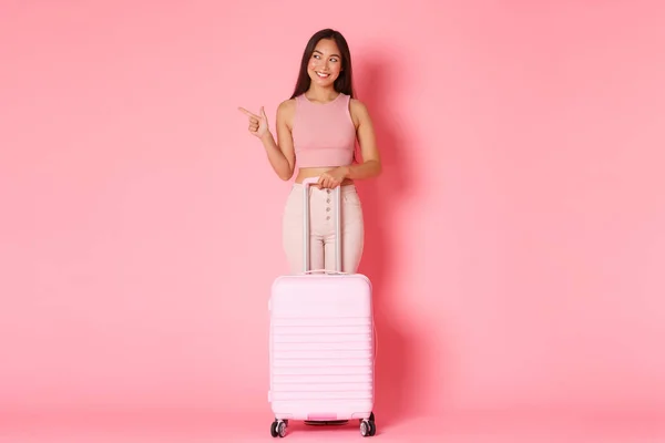 Cestování, dovolená a dovolená koncept. Plná délka potěšené atraktivní asijské dívka turista v letním oblečení, ukazující prst vlevo a při pohledu s úsměvem, stojí s kufrem — Stock fotografie