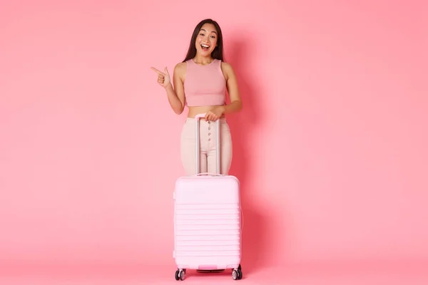 Viajar, férias e férias conceito. Comprimento completo de divertida bela menina asiática em roupas de verão, explorar países, desfrutando de verão no exterior, apontando para a esquerda, ficar com mala, fundo rosa — Fotografia de Stock