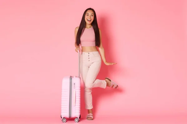 Travelling, holidays and vacation concept. Full-length of coquettish gorgeous asian girl in summer clothes, winking silly at camera, standing with suitcase, tourist packed for going abroad