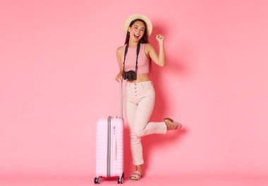 Turizm, yaz tatili, yurt dışında tatil kavramı. Neşeli Asyalı turist kız, yolculuğa hazır gezgin, bavulun yanında kamerayla ve hasır şapkayla mutlu bir şekilde zıplıyor, pembe arka plan.