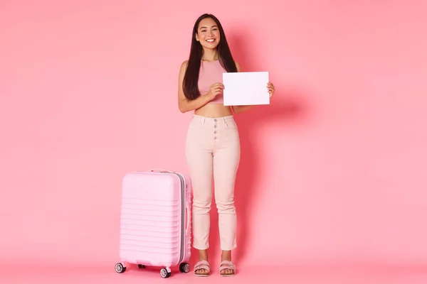 Cestování, dovolená a dovolená koncept. Plná délka krásné usměvavé asijské dívka turista s kufrem přes růžové pozadí, čeká na někoho letiště, stojící s nápisem na bílém papíru — Stock fotografie