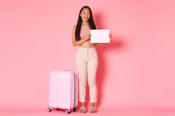 Cestování, dovolená a dovolená koncept. Plná délka koketní módní brunetka asijské dívka čeká na někoho, kdo ji vyzvednout letiště, stojící s cedulí a kufr přes růžové pozadí — Stock fotografie