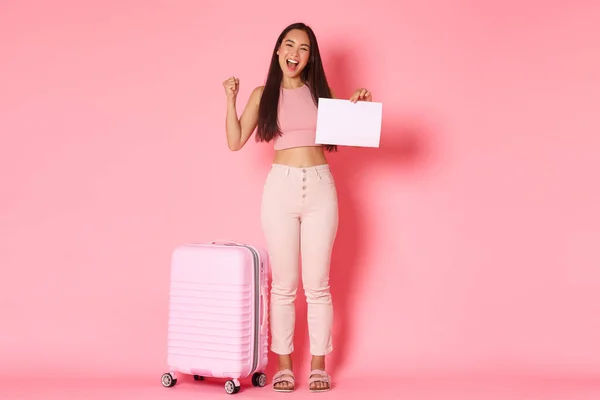 Travelling, holidays and vacation concept. Full-length of cheerful successful asian girl got opportunity travel abroad, standing with suitcase, fist pump in rejoice, holding piece of paper — Stock Photo, Image