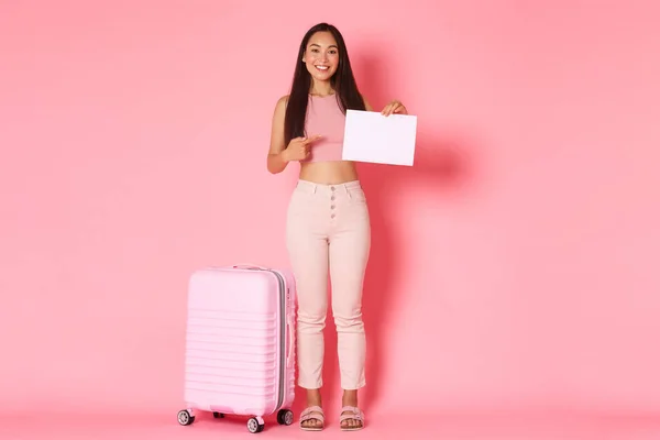 Reisen, Urlaub und Urlaubskonzept. Ganzes Lächeln attraktive asiatische Mädchen wartet auf jemanden am Flughafen, zeigt mit dem Finger auf leeres Blatt Papier mit Koffer über rosa Hintergrund — Stockfoto