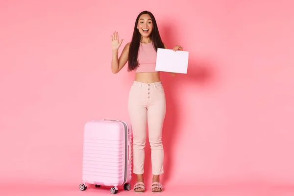 Reisen, Urlaub und Urlaubskonzept. Ganzer Länge freundlich lächelnde asiatische Mädchen, die Touristen auf dem Flughafen mit Schild treffen, leeres Papier in der Hand und winkende Hand in der Nähe von Koffer, rosa Hintergrund — Stockfoto