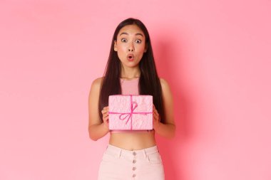 Holidays, celebration and lifestyle concept. Surprised and intrigued asian girl celebrating birthday receive wrapped gift, wonder what inside of present, standing amused over pink background clipart
