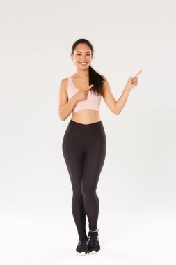 Gülümseyen, spor giyimli, zayıf Asyalı güzel bir kız, sağ üst köşeyi işaret ediyor, spor salonu üyeliği için davet ediyor, egzersiz teklifi sunuyor, fitness koçu sağlıklı diyet önerileri gösteriyor.