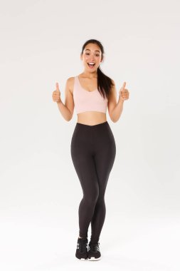 Şaşırmış ve tatmin olmuş bir şekilde gülümseyen fitness kızı, aktif giyinen bayan sporcu yeni spor salonunu ya da egzersiz programını seviyor, baş parmaklarını gösteriyor, antrenmanlara başlamaya teşvik ediyor ya da egzersiz yapıyor.