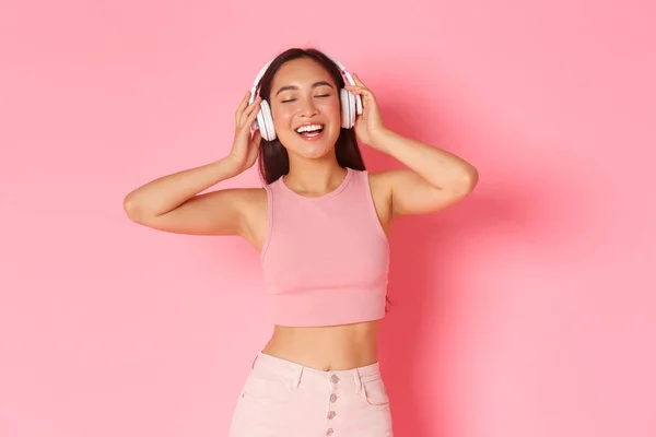 Despreocupado hermosa chica asiática en auriculares, bailando con los ojos cerrados y relajados, sonrisa feliz, disfrutando de escuchar música, escuchar canción favorita, de pie sobre fondo rosa — Foto de Stock