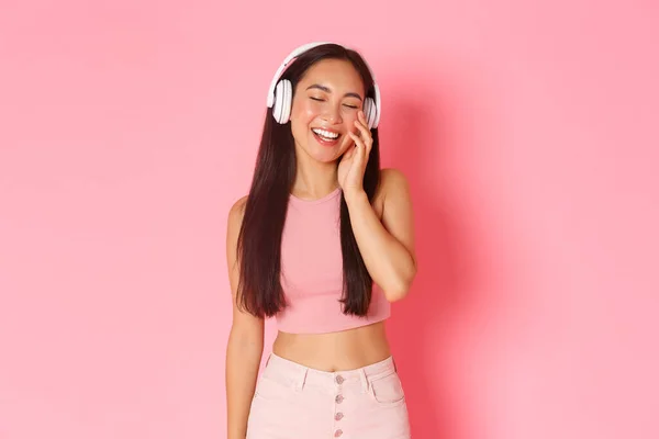 Retrato de feliz y feliz, sonriente chica asiática atractiva en auriculares inalámbricos, escuchar música o divertido podcast, cerrar los ojos alegremente, de pie sobre fondo rosa — Foto de Stock