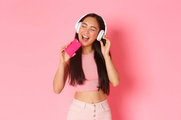 Pas-up portret atrakcyjnej stylowej azjatyckiej dziewczyny zamknij oczy i śpiewając wzdłuż piosenki podczas słuchania muzyki w słuchawkach, grając w grę karaoke na telefon komórkowy, stojąc nad różowym tle — Zdjęcie stockowe