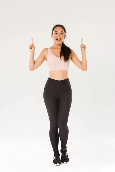 Deporte, gimnasio y concepto corporal saludable. Longitud completa de alegre sonriente chica asiática fitness, athelte femenina en ropa deportiva mostrando publicidad, señalando los dedos hacia arriba y invitando a entrenar, entrenamiento — Foto de Stock