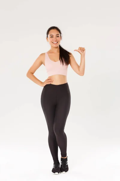 Pleine longueur de confiance souriant asiatique athelte féminine dans sportsbra et leggigns, fille de remise en forme pointant vers elle-même, mener un mode de vie actif et sain, étant professionnel, debout fond blanc — Photo