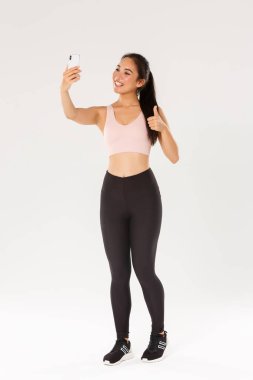 Tam boy gülümseyen kadın yaşam tarzı blogcusu, spor giyim sektöründe Asyalı fitness kızı selfie çekiyor, baş parmaklarını kaldırıyor, online takipçiler için egzersiz egzersizleri yapıyor, antrenmanlardan canlı yayın yapıyor.