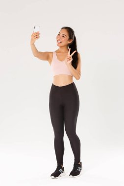 Tam boy Asyalı fitness kızı, spor giyim sportmenliği barış jesti gösteren ve cep telefonuyla selfie çeken spor salonundaki kadın spor salonunda egzersiz yapan ve antrenman sırasında kendi fotoğraflarını çeken.