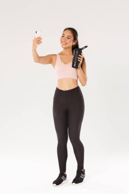 Tam boy gülümseyen ince ve sağlıklı Asyalı kız selfie fitness eğitimi alıyor, kadın blogcu ve atlet cep telefonunun kamerasında su şişesini gösteriyorlar, beyaz arka planda duruyorlar.