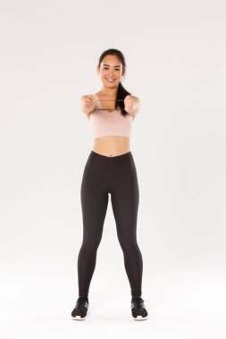 Gülümseyen, yakışıklı ve ince Asyalı fitness koçu aktif giyimli bayan sporcu egzersiz ekipmanları gösteriyor, direnç bandı tutuyor, egzersiz yapıyor, beyaz arka plan