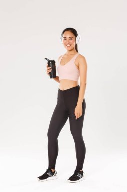 Tam boy gülümseyen ince ve mutlu fitness kızı, spor salonunda kulaklıkla çalışan Asyalı bayan sporcu, antrenman sırasında müzik dinleyen, egzersizler arasında şişeden su içen, beyaz arka plan