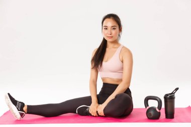 Antrenmandan önce lastik mindere uzanan, kettlebell ile antrenman yapan spor salonunda yeterince su içen, spor yaparken beyaz arka planda duran, aktif giyimli Asyalı fitness kızı portresi.