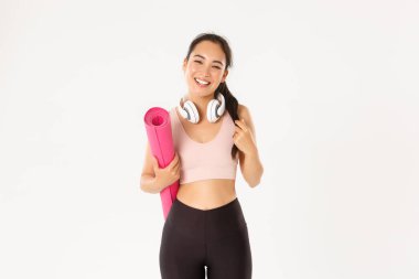 Spor, refah ve aktif yaşam tarzı konsepti. Gülümseyen mutlu Asyalı fitness kızı kulaklıklar ve spor kıyafetlerle antrenman veya yoga için lastik minder taşır, beyaz arka planda kaygısızca güler.