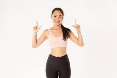 Spor, refah ve aktif yaşam tarzı konsepti. Gülümseyen ince ve tatlı Asyalı fitness kızı, parmaklarını gösteren bayan sporcu, egzersiz malzemeleri satış promosyonu veya spor salonu üyeliği teklifi