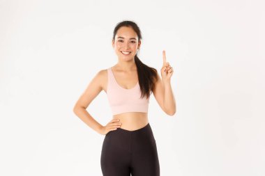 Spor, refah ve aktif yaşam tarzı konsepti. Gülümseyen Asyalı fitness eğitmeni, spor giyim dersindeki kız, birinci kuralı gösteriyor, parmağını kaldır ve gülümse, beyaz arka planda dur.