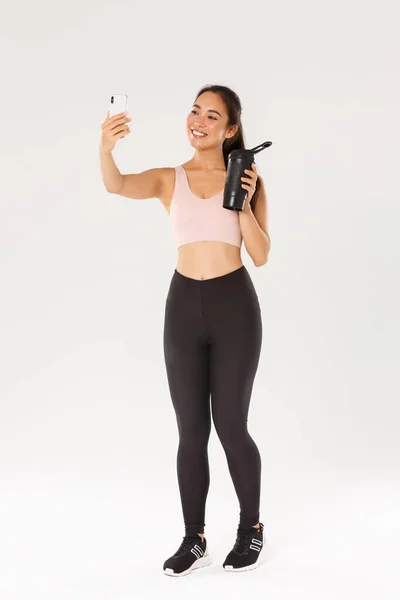 Longitud completa de la sonrisa delgada y saludable chica asiática tomando sesión de entrenamiento de fitness selfie, blogger femenina y athelte mostrando botella de agua en la cámara del teléfono móvil, de pie sobre fondo blanco — Foto de Stock