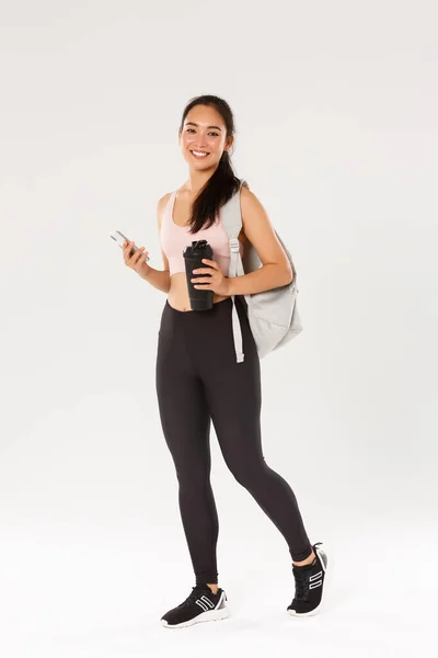 Longitud completa de sonreír sano, delgado asiático chica ir entrenamiento de fitness, hembra athelte llevar mochila con equipo de entrenamiento y botella de agua, el uso de la aplicación de deportes de teléfono móvil, fondo blanco — Foto de Stock