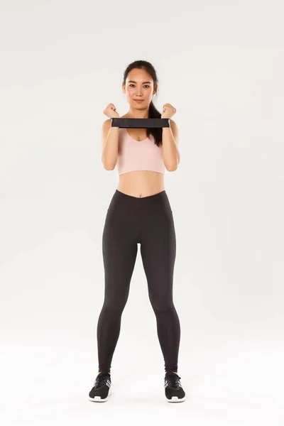 Comprimento total de sorrir e determinado, morena magro asiático fitness girl, treinador de treinamento mostrando exercícios com resistência esticando corda, olhando para a câmera, demonstrar equipamento de treino — Fotografia de Stock