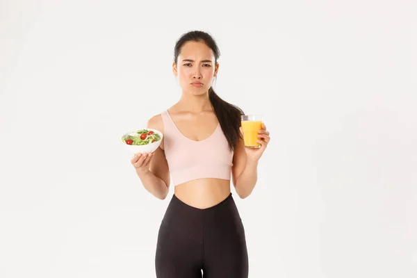 Deporte, bienestar y concepto de estilo de vida activo. Sulking y renuente linda chica asiática odia comer sano, la celebración de ensalada y jugo de naranja con molesta cara de puchero, odio estar en la dieta y el entrenamiento —  Fotos de Stock