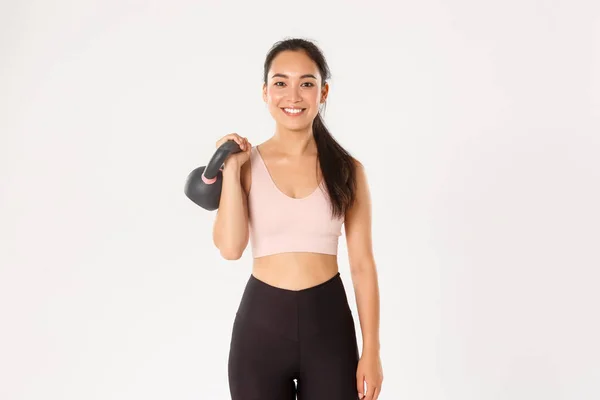 Esporte, bem-estar e conceito de estilo de vida ativo. Sorrindo forte e magro asiático fitness menina, atleta feminino segurando kettlebell e olhando despreocupado, ganhando músculos no ginásio, de pé fundo branco — Fotografia de Stock