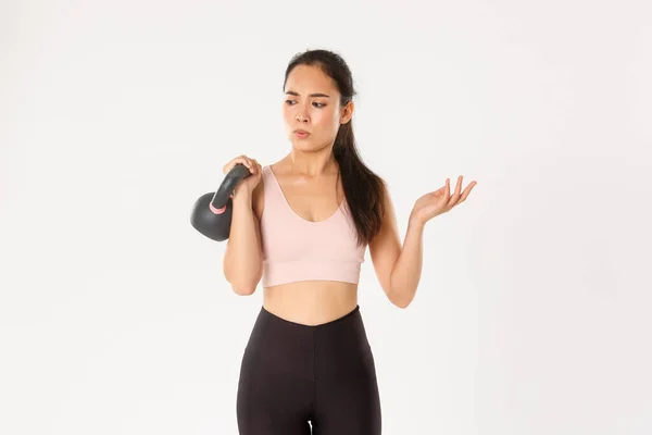Sport, wellbeing and active lifestyle concept. Confused asian fitness girl in activewear looking puzzled, lifting kettlebell, getting strong with workout and healthy diet, standing white background — Stock Photo, Image