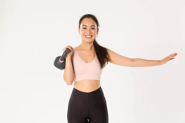Αθλητισμός, ευεξία και ενεργός τρόπος ζωής. Χαμογελώντας όμορφη ασιατική fitness girl, γυμναστής επεκτείνει ένα χέρι και να άρει kettlebell, bodybuilding, κερδίζοντας μυϊκή δύναμη, στέκεται λευκό φόντο — Φωτογραφία Αρχείου