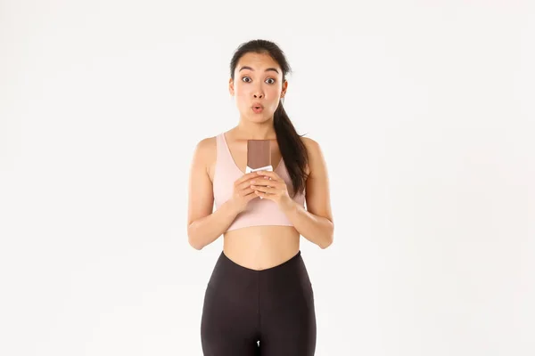 Sport, wellbeing and active lifestyle concept. Amused and wondered asian female athlete holding chocolate protein bad and looking excited, eating healthy sweets for prolonged workout — Stock Photo, Image