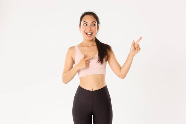 Sport, wellbeing and active lifestyle concept. Excited slim and cute asian fitness girl, female athlete pointing fingers upper right corner, showing workout equipment sale promo or gym offer