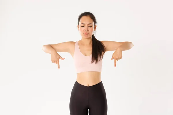 Deporte, bienestar y concepto de estilo de vida activo. Enfurruñada y decepcionada chica de entrenamiento asiática, atleta femenina en ropa deportiva frunciendo el ceño molesto, apuntando y mirando hacia abajo a lo malo, fondo blanco — Foto de Stock