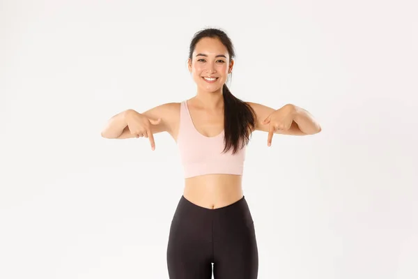 Esporte, bem-estar e conceito de estilo de vida ativo. Sorrindo ajuste e magro feminino instrutor de fitness, asiático treino menina no ginásio e mostrando algo, apontando os dedos para baixo, de pé fundo branco — Fotografia de Stock