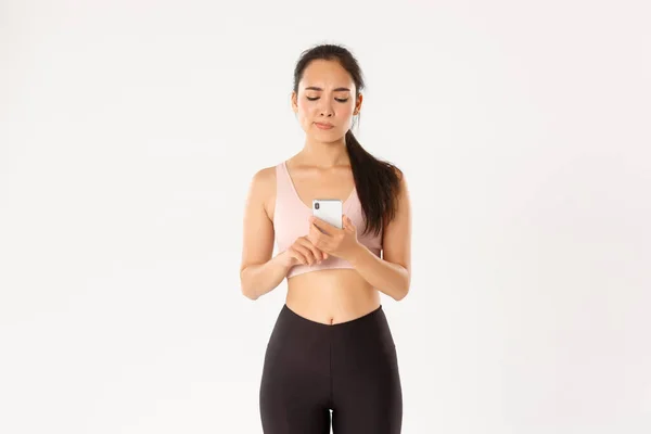 Sport, technology and active lifestyle concept. Fitness girl looking at her smartphone screen with upset face and smirk, disappointed with running results of workout tracker application — Stock Photo, Image