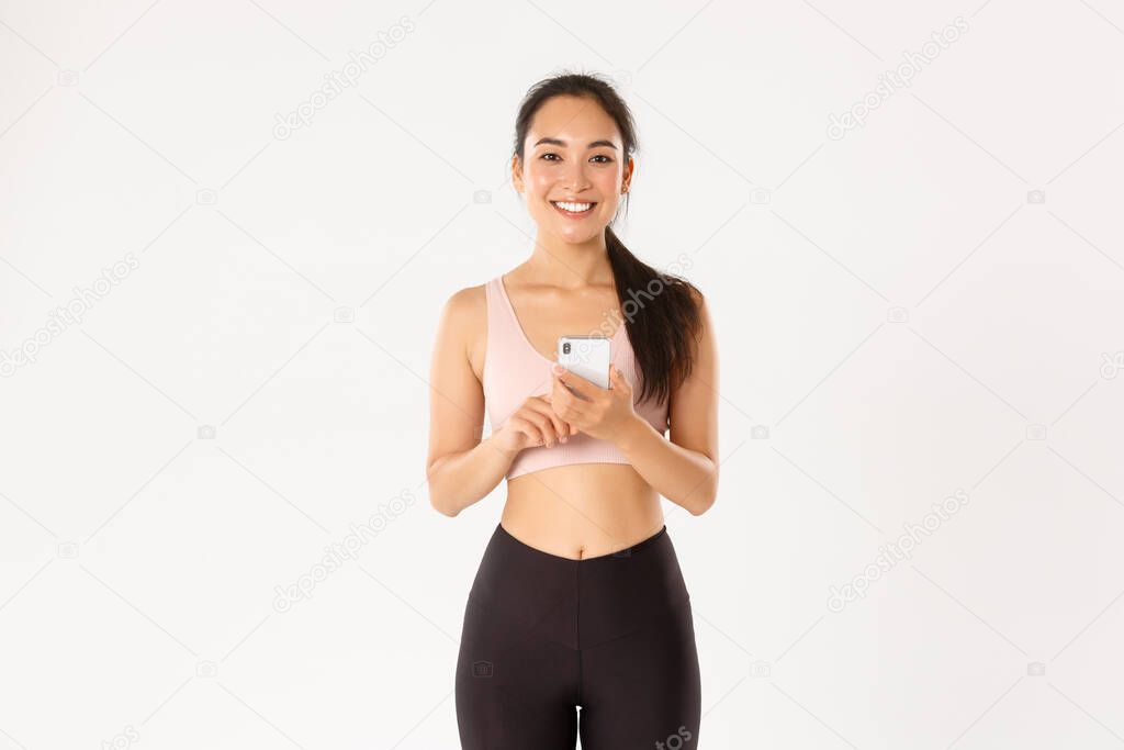 Sport, technology and active lifestyle concept. Smiling satisfied asian fitness girl, sportswoman holding smartphone, using running tracker app, checking heartrate during workout, white background