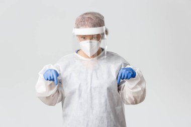 Tıp çalışanları, covid-19 salgını, koronavirüs konsepti. Etkilenmiş ve meraklı kadın doktor, kişisel koruma ekipmanlarında çalışan doktor ilgi çekici ve parmaklarını aşağıya doğru gösteren gri arka plan