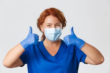 Tıp çalışanları, covid-19 salgını, koronavirüs konsepti. Yakın plan gülümseyen kızıl saçlı kadın doktor, önlüklü hemşire, tıbbi maske ve eldivenler.