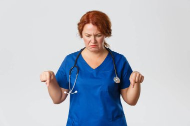 Tıp, sağlık ve koronavirüs konsepti. Hüsrana uğramış ve hayal kırıklığına uğramış orta yaşlı kadın doktor, tiksinti ve tiksintiyle bakan hemşire, yüzünü buruşturan, parmaklarını aşağıya doğru gösteren.