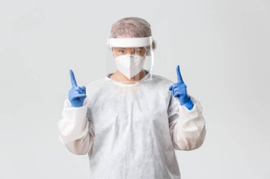 Tıp çalışanları, covid-19 salgını, koronavirüs konsepti. Etkilenmiş ve meraklı kadın doktor, kişisel koruma ekipmanlarında çalışan doktor ilgi çekici ve işaret eden gri arka plan