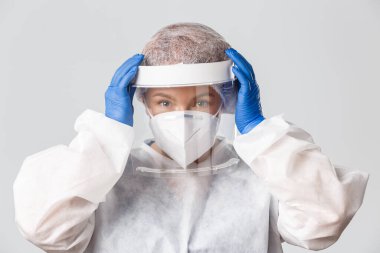 Tıp çalışanları, covid-19 salgını, koronavirüs konsepti. Umutlu gülümseyen kadın doktor hasta insanlarla bulaşıcı bölgeye girmeden önce kişisel koruyucu giysilerini giydi ve yüz kalkanını tamir etti.