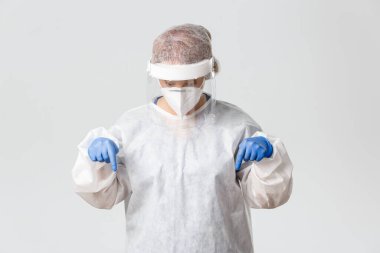 Tıp çalışanları, covid-19 salgını, koronavirüs konsepti. Şahsi koruma ekipmanlarıyla etkileyici ve şaşkın bir kadın doktor sersemlemiş görünüyor, parmaklarını aşağıya doğru gösteriyor, afiş okuyor, arka planda gri renkler var.