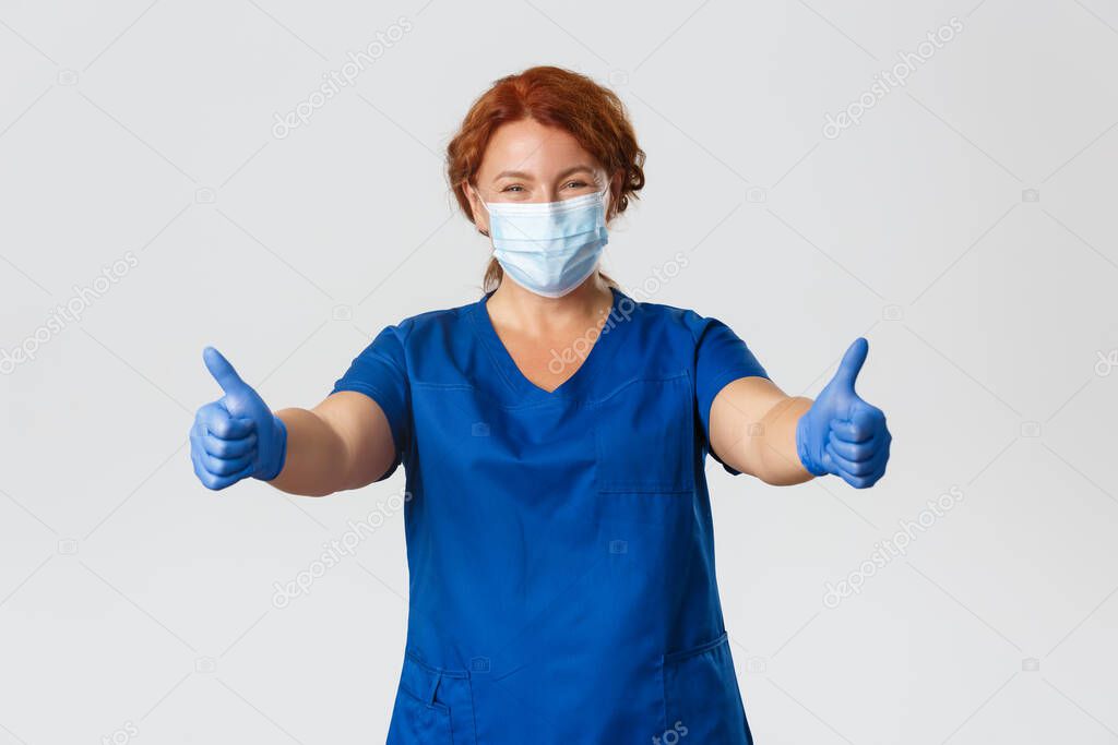 Medical workers, covid-19 pandemic, coronavirus concept. Happy smiling female doctor, middle-aged nurse in medical mask and gloves showing thumbs-up, gurantee quality service at clinic