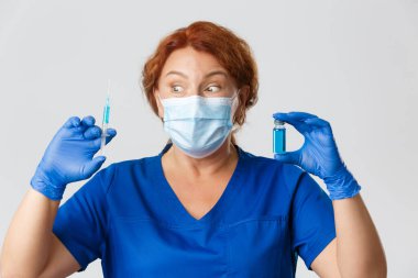 Tıp çalışanları, covid-19 salgını, koronavirüs konsepti. Şaşırmış ve heyecanlı yüz maskeli kadın doktor ve eldivenler sonunda virüsten aşılar elde ettiler, elinde heyecan verici bir ifade olan ampul ve şırınga vardı.