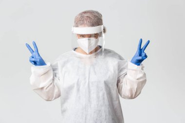 Tıp çalışanları, covid-19 salgını, koronavirüs konsepti. Kişisel koruma ekipmanlarında neşeli, gülümseyen ve iyimser bayan doktor barış jestleri gösteriyor, hastaları tedavi ediyor, pozitif kalın.