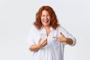 Teknoloji ve iletişim konsepti. Kaygısız, mutlu, orta yaşlı, mutlu bir kadın. Akıllı telefonu işaret ederken gülümsüyor. Harika bir uygulama öneriyor.